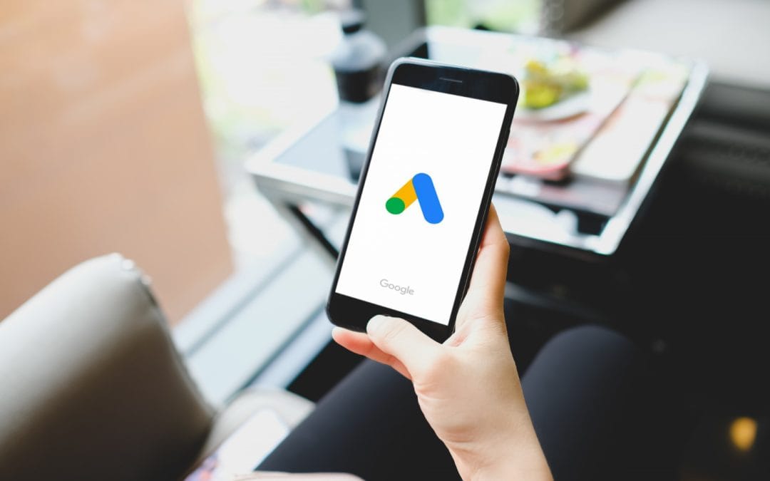 A person holding a smartphone displaying the Google Ads app logo on the screen.